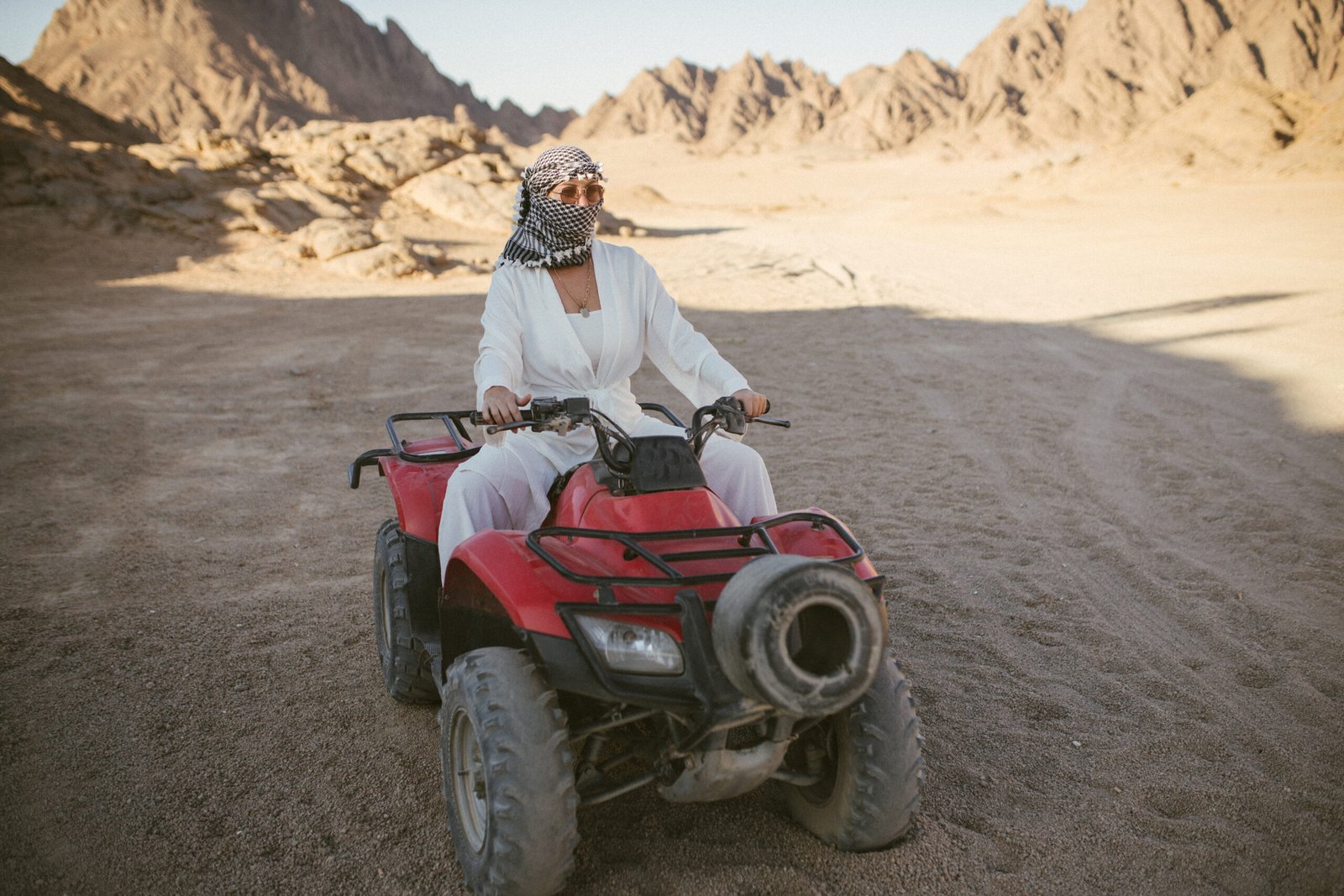 A rather attractive woman is going on a safari, driving an ATV in the desert. Beautiful young woman with a scarf on her head sits on a red four-wheeled ATV and rides towards adventure.